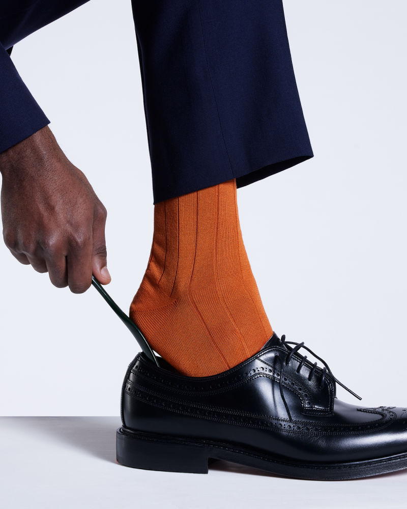 a male models lower leg wearing orange socks, using a shoe horn to put on a pair of black brogues