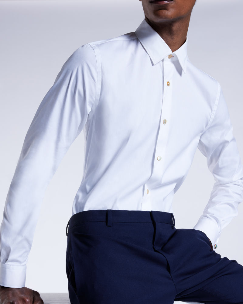 a male model sat on a white box, wearing a white shirt and navy suit trousers