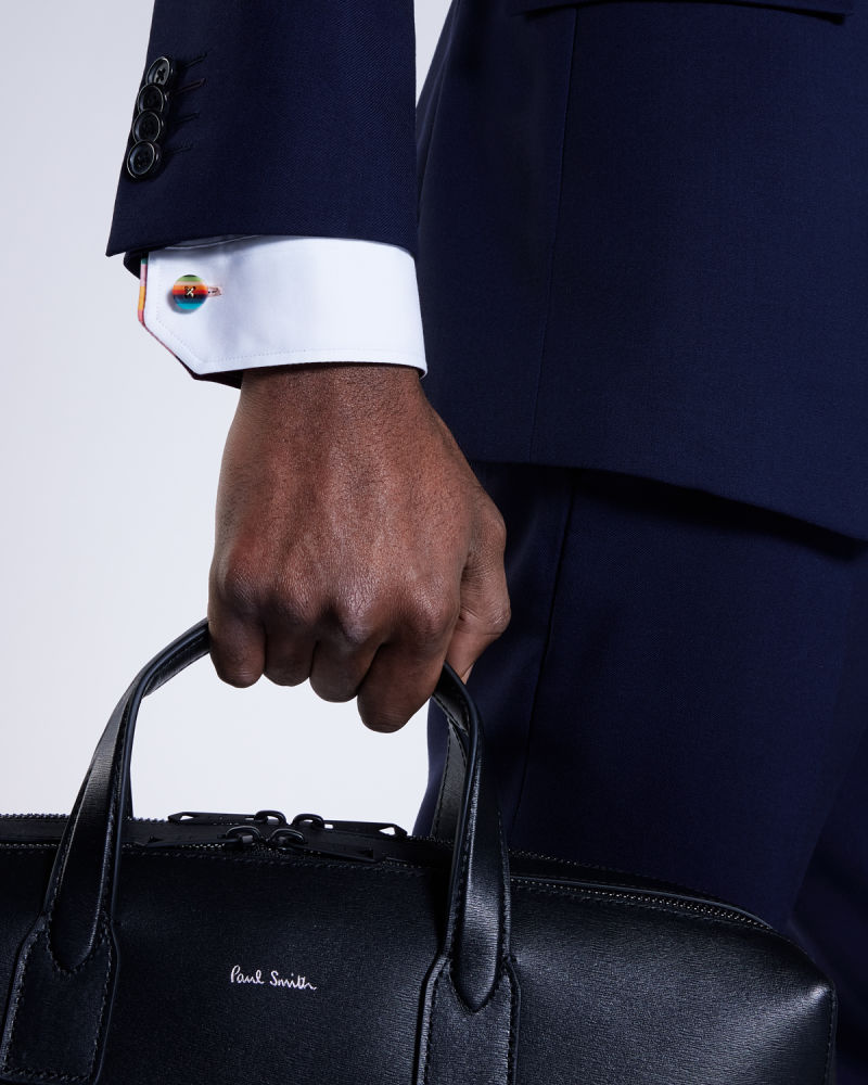 a male models arm wearing a colourful striped button cufflink, with a navy suit holding a black leather business bag