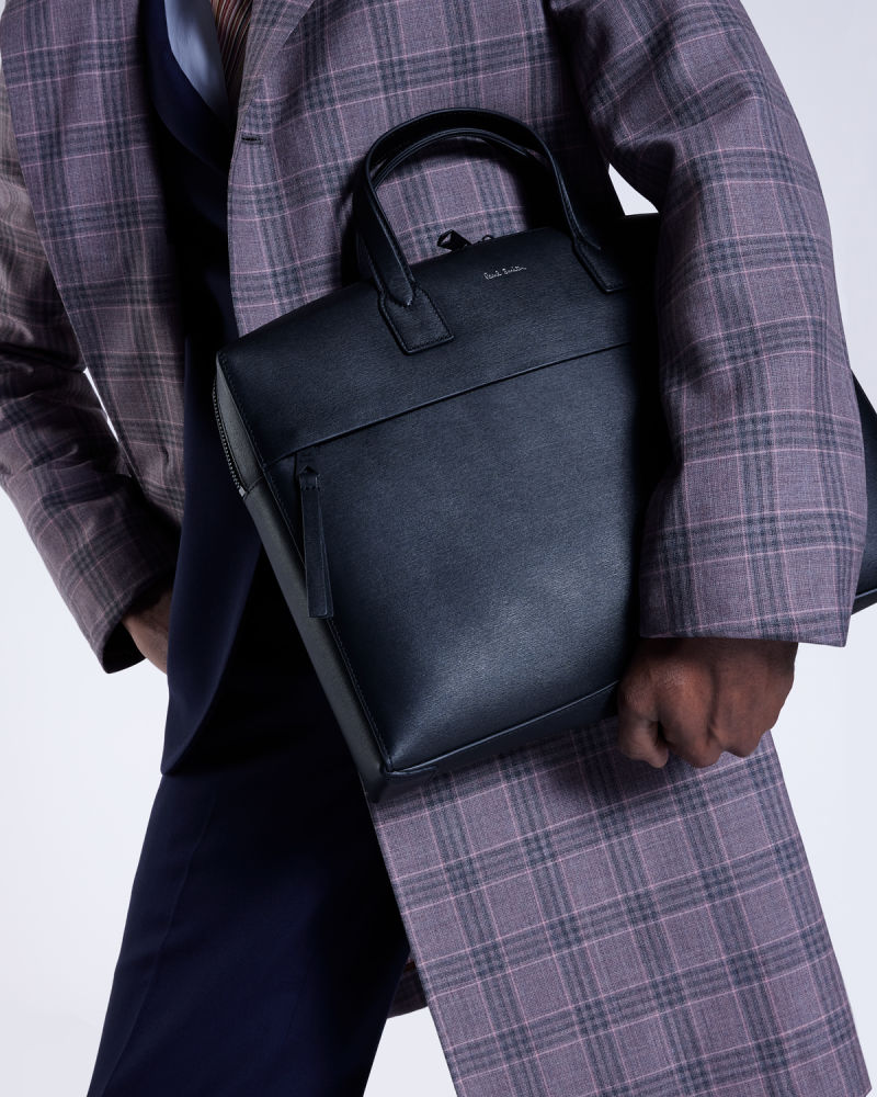 a male model wearing a navy suit with a long checked overcoat, holding a black leather business bag
